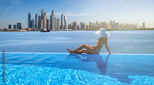 Canvas Print Beautiful panorama of Dubai Marina skyline in a background with a pool, deck chair and woman with a white hat