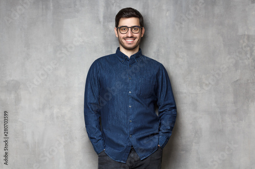 Young handsome man wering eyeglasses, smiling and feeling confident, standing against gray textured wall background