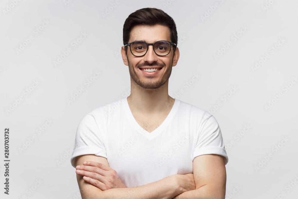 Fototapeta premium Portrait of smiling young man in casual white t-shirt, looking at camera, standing with crossed arms isolated on gray background