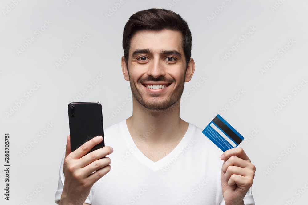 Studio portrait of young smiling man wearing white t-shirt, holding credit card and phone, isolated on gray background Studio portrait of young smiling man wearing white t-shirt, holding credit card and phone, isolated on gray background