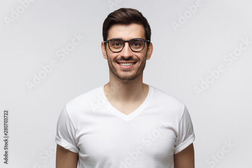 Eyewear fashion. Portrait of smiling handsome young man in casual white t-shirt and trendy glasses, looking at camera, isolated on gray background © Damir Khabirov
