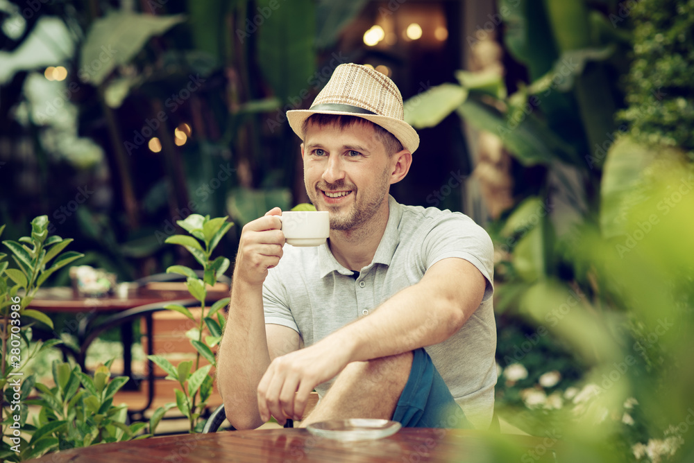 Young man in sunhat sits in cafe outdoors, drinks coffee during lunch time, rests, enjoys drink.