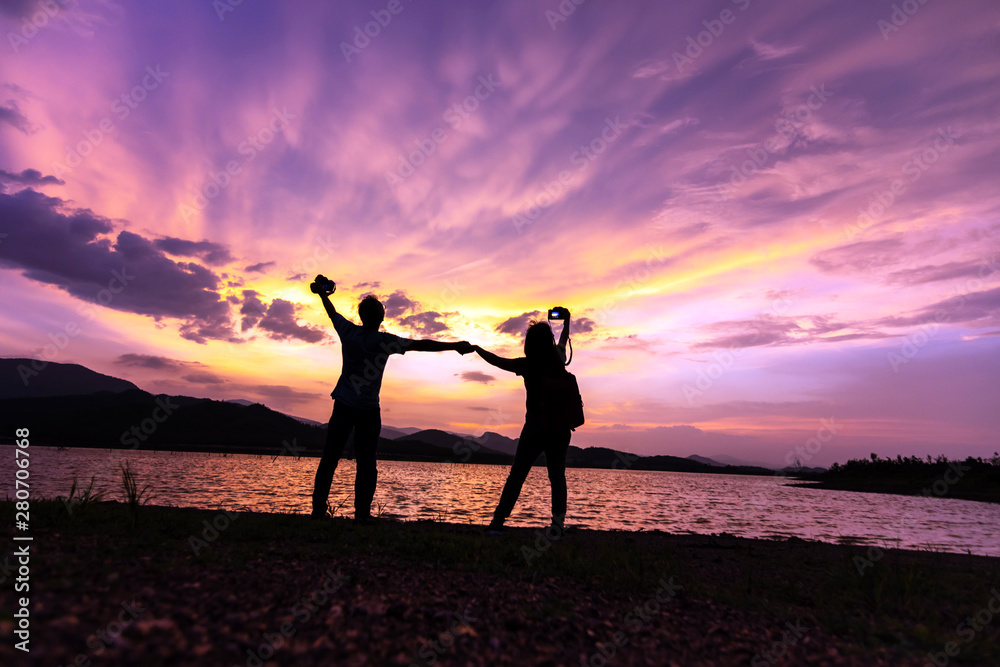 Silhouette of couple holding hands and take a photo by camera while dramatic sunset sky with beautiful clouds. Traveling and vacation concept.