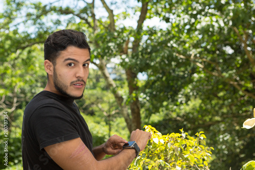 Young latin man portrait indoors next to a window with nature as background. He is smiling He is using the smartwatch. his is wearing.