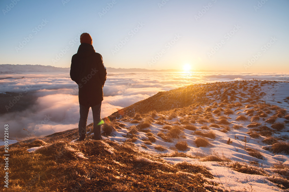 View From the back. A lonely standing man high in the mountains looks at the setting sun and the sunset horizon with a valley filled with clouds. The concept of tourism travel and male loneliness View From the back. A lonely standing man high in the mountains looks at the setting sun and the sunset horizon with a valley filled with clouds. The concept of tourism travel and male loneliness