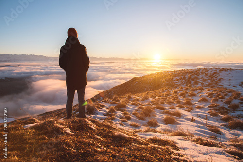 View From the back. A lonely standing man high in the mountains looks at the setting sun and the sunset horizon with a valley filled with clouds. The concept of tourism travel and male loneliness