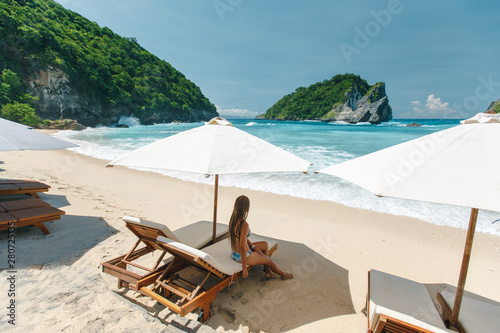 Bild auf Leinwand Youn woman in bikini  resting under umbrella facing the seaside in a deserted beach with deep blue sky and enjoying the summer