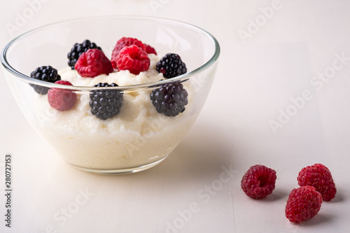 Semolina pudding porridge dessert with  blackberry and raspberry in glass bowl near with scattered heap berries on white wooden background, breakfast, isolated
