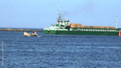 RIGA/LATVIA - JUNE 04 2019: Motorboat speeds past huge green vessel with large containers sailing along river surface against boundless blue sky on June 04 in Riga