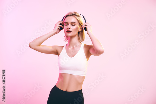 My favourite playlist. Cute and sporty woman adjusting headphones and keeping eyes closed while standing against pink background © Friends Stock