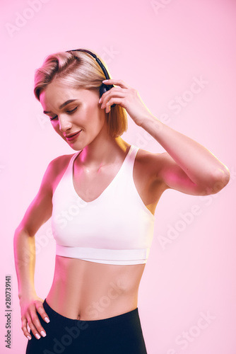 Music making me happy. Cute and sporty woman adjusting headphones and keeping eyes closed while standing against pink background. © Friends Stock