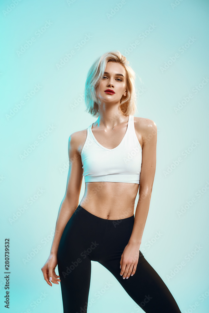 Beauty portrait. Sexy young woman in white top and black leggings looking  at camera while standing against blue background in studio Stock Photo |  Adobe Stock