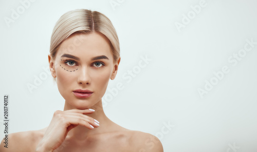 Perforation lines. Beautiful and young blonde woman with black surgical lines on eyelids and under eyes and looking at camera while standing against grey background photo