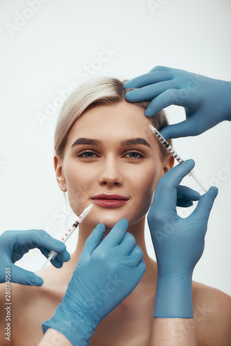 Creating beauty. Portrait of young pretty woman looking at camera and smiling while doctors in blue medical gloves making injections in her face