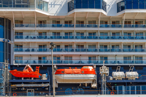 There are two orange lifeboats on the board of cruise ship. A eight floors cruise liner with orange lifeboats is in the port in Koper, Slovenia