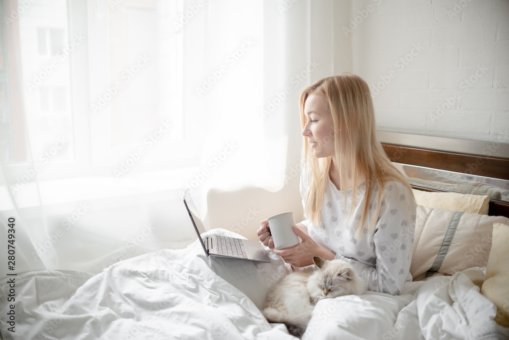 Work from home concept . Blonde Giirl typing and using laptop. Hygge with fluffy cat, pajama and coffee cup. Horizontal copyspace Work from home concept . Blonde Giirl typing and using laptop. Hygge with fluffy cat, pajama and coffee cup. Horizontal copyspace