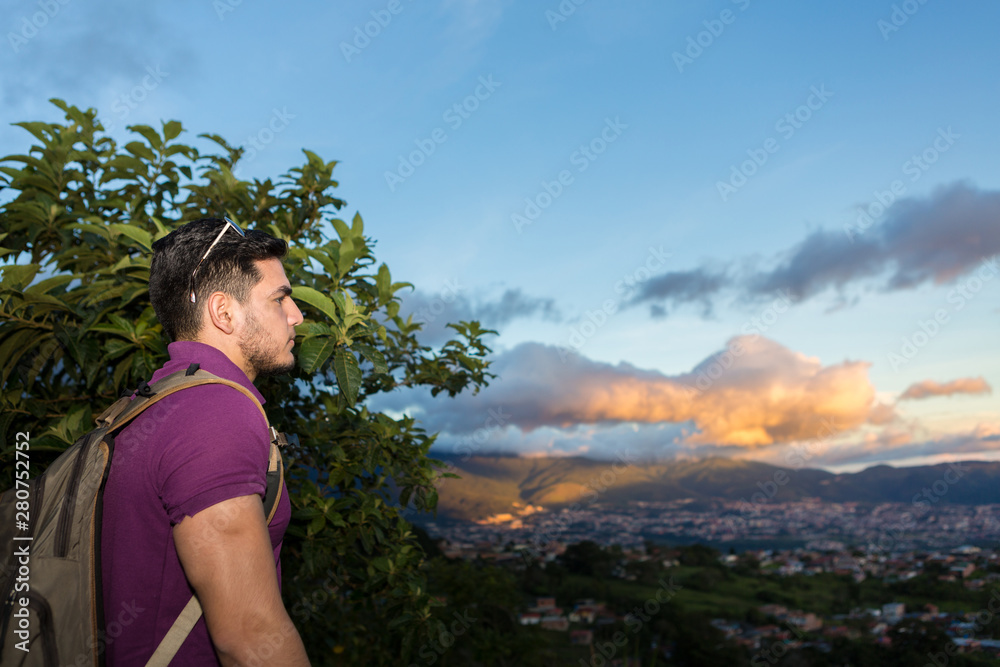 Young Man enjoys the scene of nature during sunset at the mountains looking down to the city. He is hiking with a backpack. 