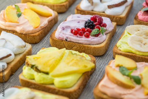Assorted toasts with fruits and berries on wipped cream on a wood board on light background. Summer traditional homemade dessert.
