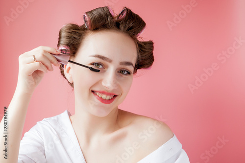 nice cheerful young the girl in the curler paints an eyebrow on pink background