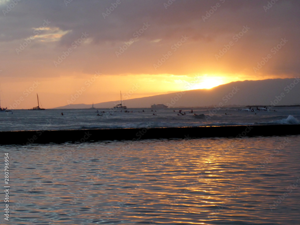 Sunsets behind Waianae mountains with light reflecting on ocean and illuminating the sky with surfers and boats in the water off Waikiki Sunsets behind Waianae mountains with light reflecting on ocean and illuminating the sky with surfers and boats in the water off Waikiki