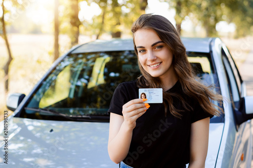 Attractive young woman wearing black T-shirt standing in front of modern car bragging about receiving driving license showing licence to the camera and feeling happy photo