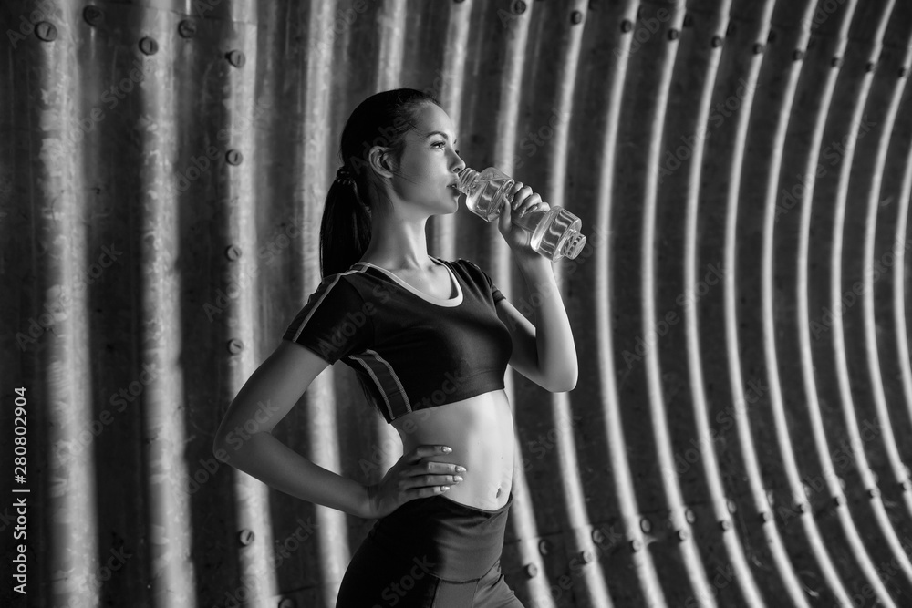 Obraz premium Beautiful slim brunette woman drinking water from bottle after running. Fitness young model working out outdoor on tunnel urban gray background. Black and white.
