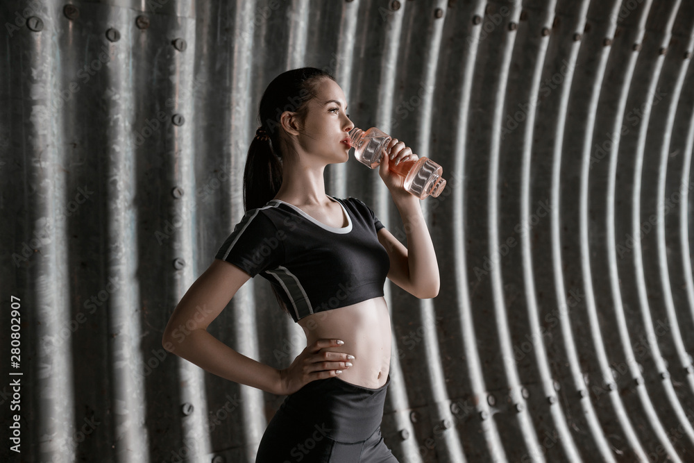 Obraz premium Beautiful slim brunette woman drinking water from bottle after running. Fitness young model working out outdoor on tunnel urban gray background.