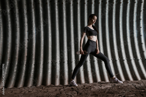 Sports woman in fashion sportswear on tunnel urban gray background. Fitness model working out outdoor. Young beautiful slim brunette girl doing warmup exercise with dumbbells.