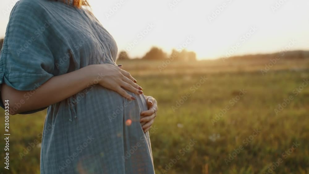 Pregnant woman dressed in linen dress and straw hat standing in meadow during sunset. Young mother to be feels happy, smiles into camera looks at sun, stroking and touching pregnant belly with hands