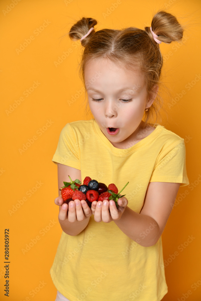 Baby girl kid is much surprised with a big handful of strawberry raspberry blueberry cherry in her palms on yellow 