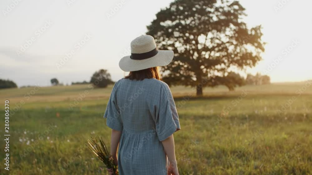 Pregnant woman walking in meadow during sunset. Pretty mother to be dressed in linen dress and straw hat with flower bouquet in the field by oak tree takes pleasure and turns to camera and smiles Pregnant woman walking in meadow during sunset. Pretty mother to be dressed in linen dress and straw hat with flower bouquet in the field by oak tree takes pleasure and turns to camera and smiles