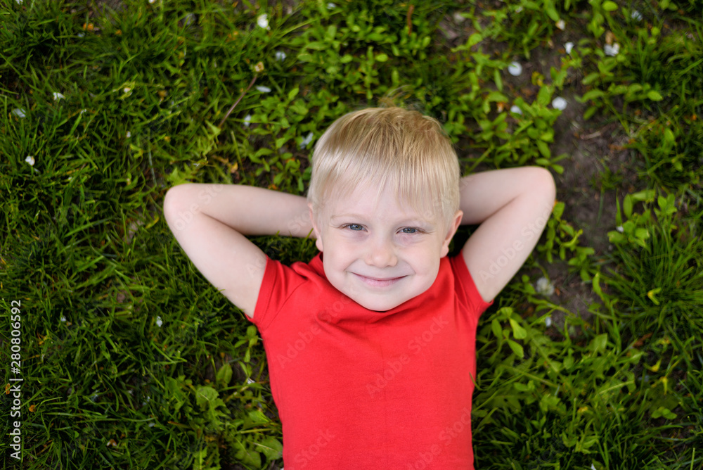Portrait of a smiling blond boy lying on the grass. Hands behind head. Rest at nature