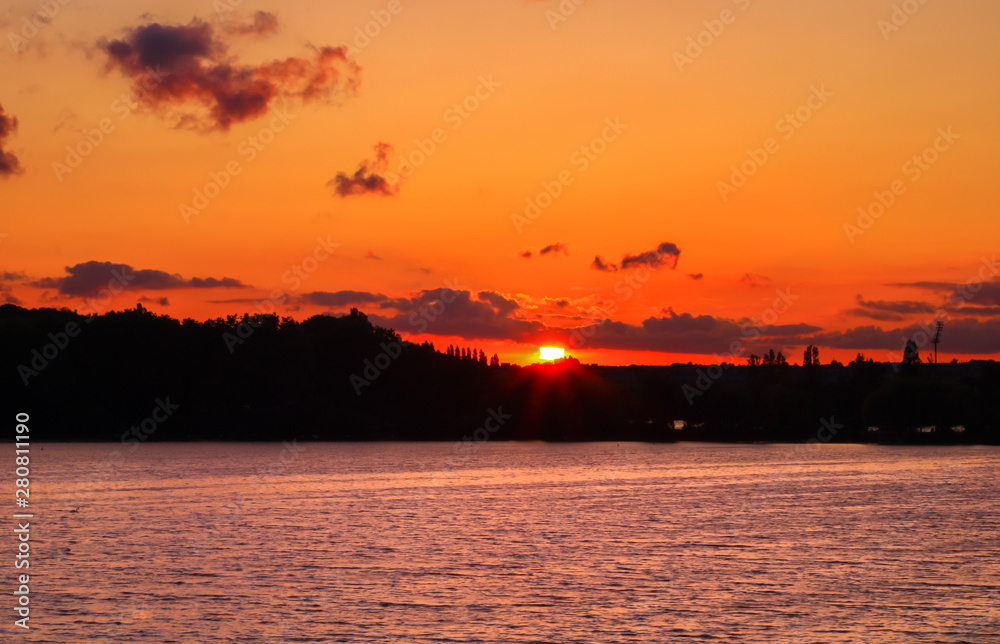 Bright sunset in an orange landscape. Half of the sun setting on the horizon between the clouds. Golden reflections on the water and silhouette of trees on a hill. Bright sunset in an orange landscape. Half of the sun setting on the horizon between the clouds. Golden reflections on the water and silhouette of trees on a hill.