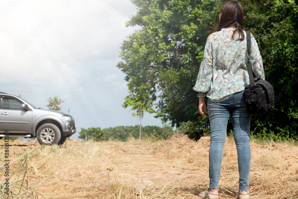 Women sitting on the dry grass with  hanging bag. Looking at the front with big trees and cars in front.