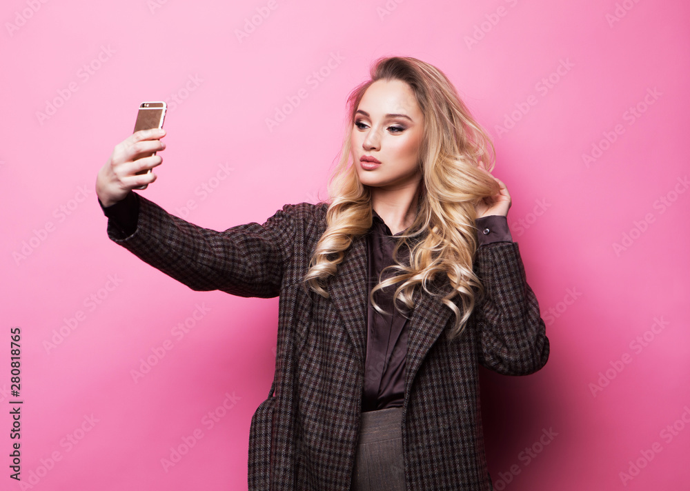 Blond girl taking photo makes self portrait on smartphone over pink background Blond girl taking photo makes self portrait on smartphone over pink background