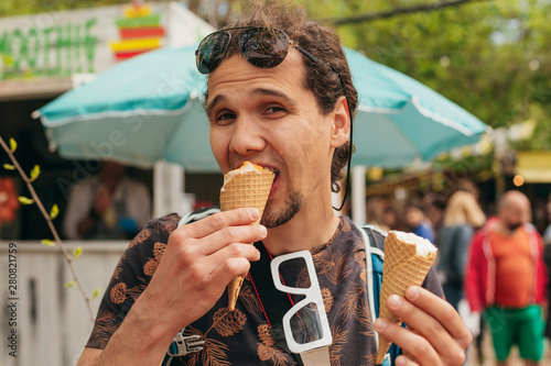 Close up portrait of a handsome young man biting two ice cream cones over fair background. Photographer with sunglasses and backpack.