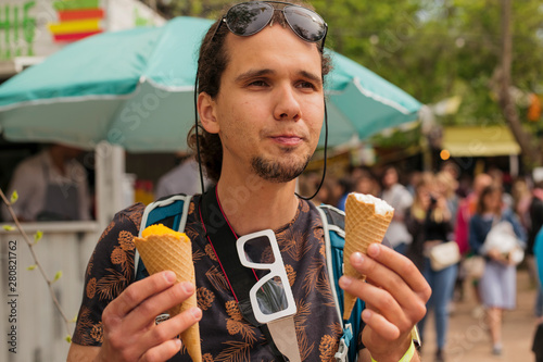 Close up portrait of a handsome young man savouring two ice cream cones over fair background. Photographer with sunglasses and backpack.
