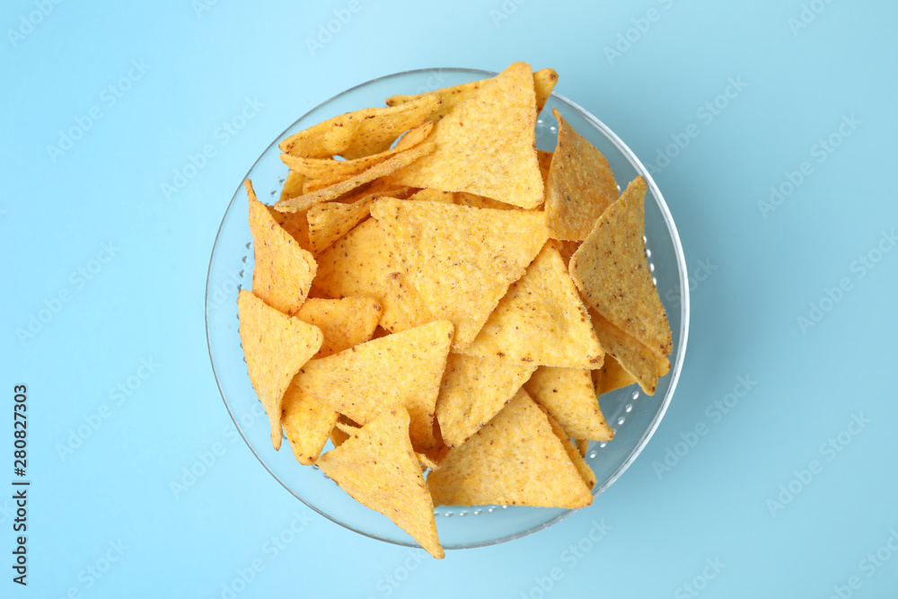 Glass bowl of tasty Mexican nachos chips on light blue background, top view