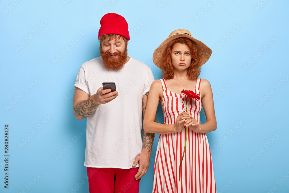 Obraz premium Unhappy redhead woman gets bored while boyfriend uses cell phone, addicted from modern technologies. Young couple have date, isolated on blue background. Bearded man with phone ignores girlfriend