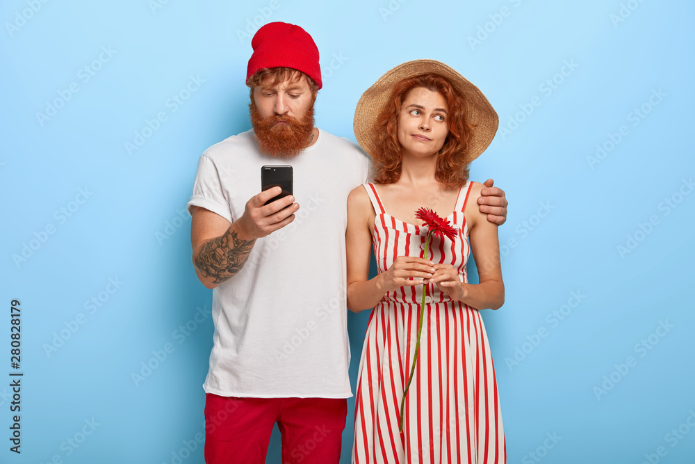 Bearded ginger man ignores wife, embraces her, chats online on mobile phone, bored woman holds red flower, looks aside with dissatisfaction, needs live communication. People, addiction, relationships