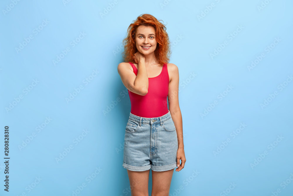 Studio shot of pretty ginger female model touches neck gently, looks with happy expression straightly at camera, wears summer fashionable outfit isolated on blue background. Feminity, emotions concept Studio shot of pretty ginger female model touches neck gently, looks with happy expression straightly at camera, wears summer fashionable outfit isolated on blue background. Feminity, emotions concept
