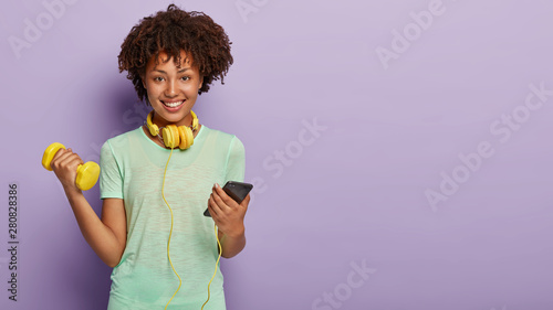 Work on yourself if you want be strong and fit. Glad Afro American woman gives motivation for sport, raises arm with dumbbell, listens audio in smart phone, invites to join sport club exercises in gym