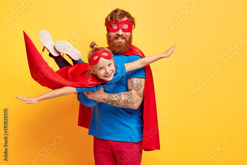 Small kid plays superman, being on fathers hands, pretends flying, spreads arms sideways, wears red mask and cloak, enjoys spare time with dad, isolated on yellow wall. Daddy hero holds daughter