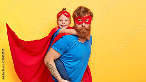 Serious bearded ginger father superhero carries small female kid on back, do good things together, wear costumes of superman, ready for children party, isolated on yellow wall. We are strong heroes