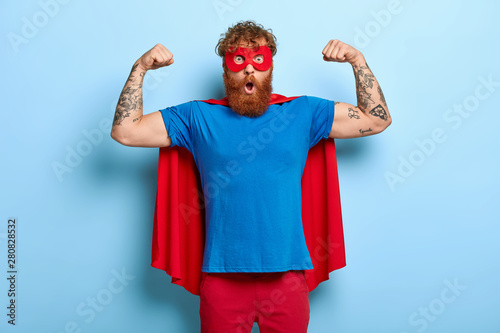 Confident red head male superhero wears mask and red cape, raises arms, shows muscles, represents power and courage, ready to defend people, has shocked expression, isolated on blue background