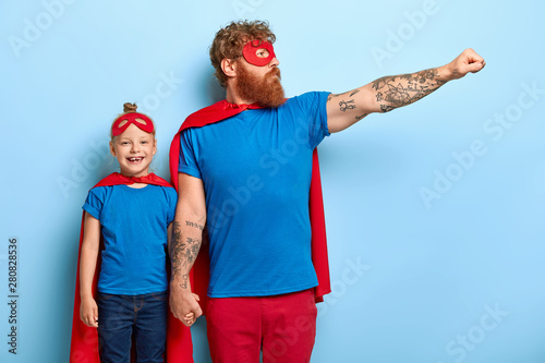 Horizontal shot of feraless strong ginger father holds hand on small kid, stretches arm and shows flying gesture, wears superhero costume, defends daughter and other people, play game together photo