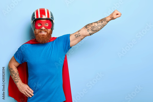 Positive bearded man has cheerful expression, keeps arm outstretched, clenches fist, ready for flight, dressed in helmet blue t shirt and cape imagines being superman uses special power to help people photo