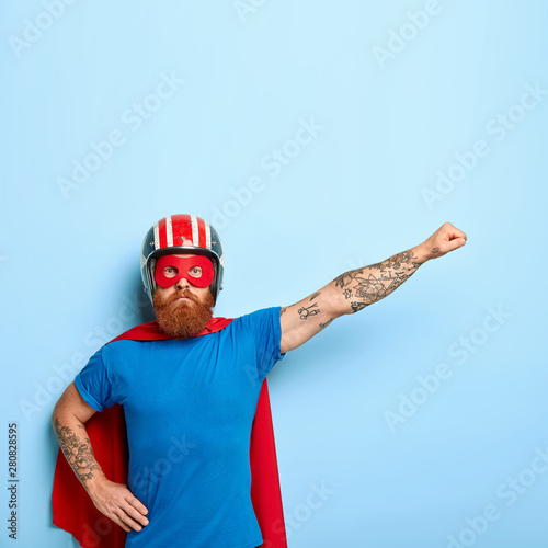 Serious confident superman pretends to fly, wears superhero costume, protective helmet, has tattooed arm, ginger beard, demonstrates power, isolated on blue wall. Leadership and strength concept photo