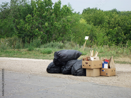 Black plastic bags of garbage and cardboard boxes with folded cardboard lie along the road. Garbage collection in the village.
