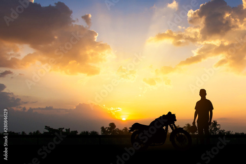 Silhouette of Back biker or rider,motorbike parking with sunset background .Traveller man standing and holding helmet beside motorcycle.Trip and lifestyle of motorbike concept,copy space.